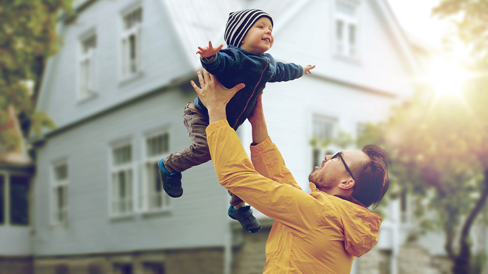 Bausparen: Vater wirft seinen Sohn vor einem Haus in die Luft.
