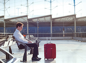 Dienstreiseversicherung: Geschäftsmann sitzt mit seinem Koffer in der Abflughalle