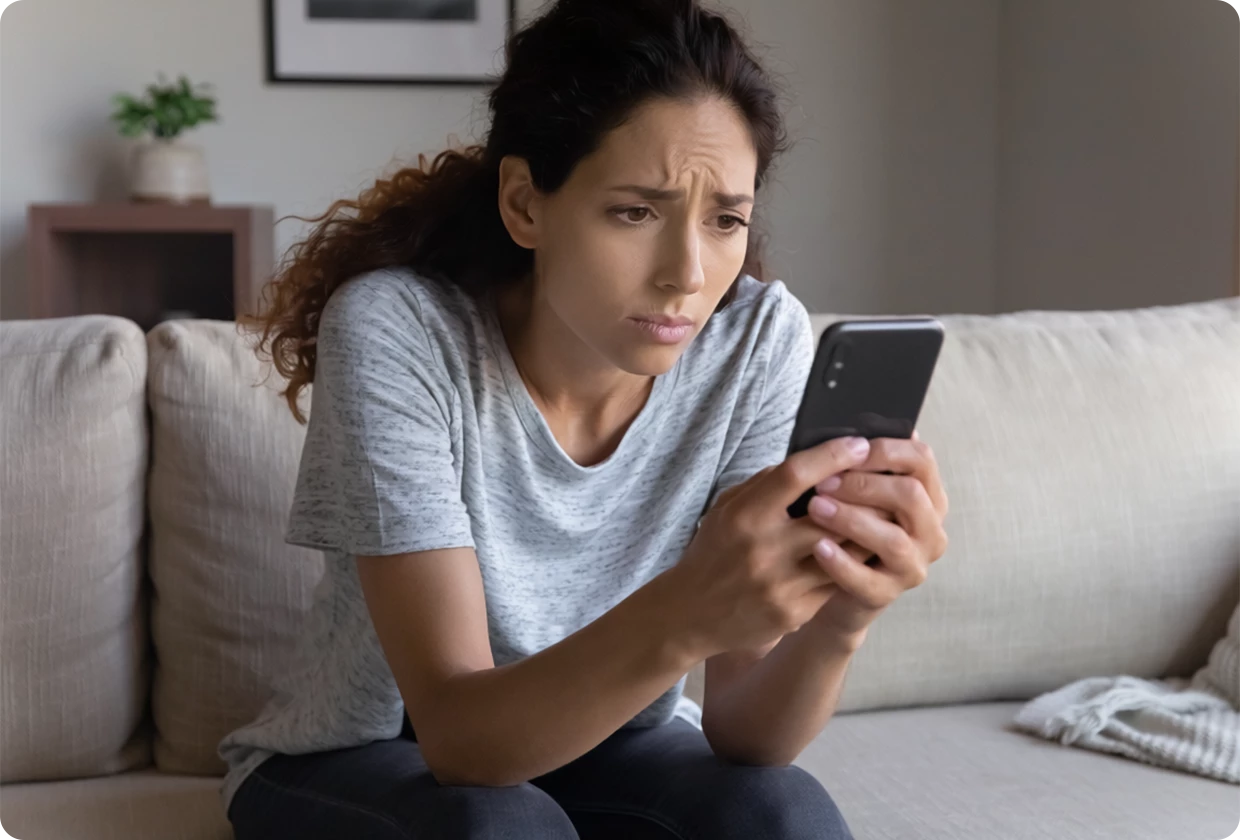 Junge Frau, die auf einer Couch sitzt und besorgt auf ihr Smartphone schaut.