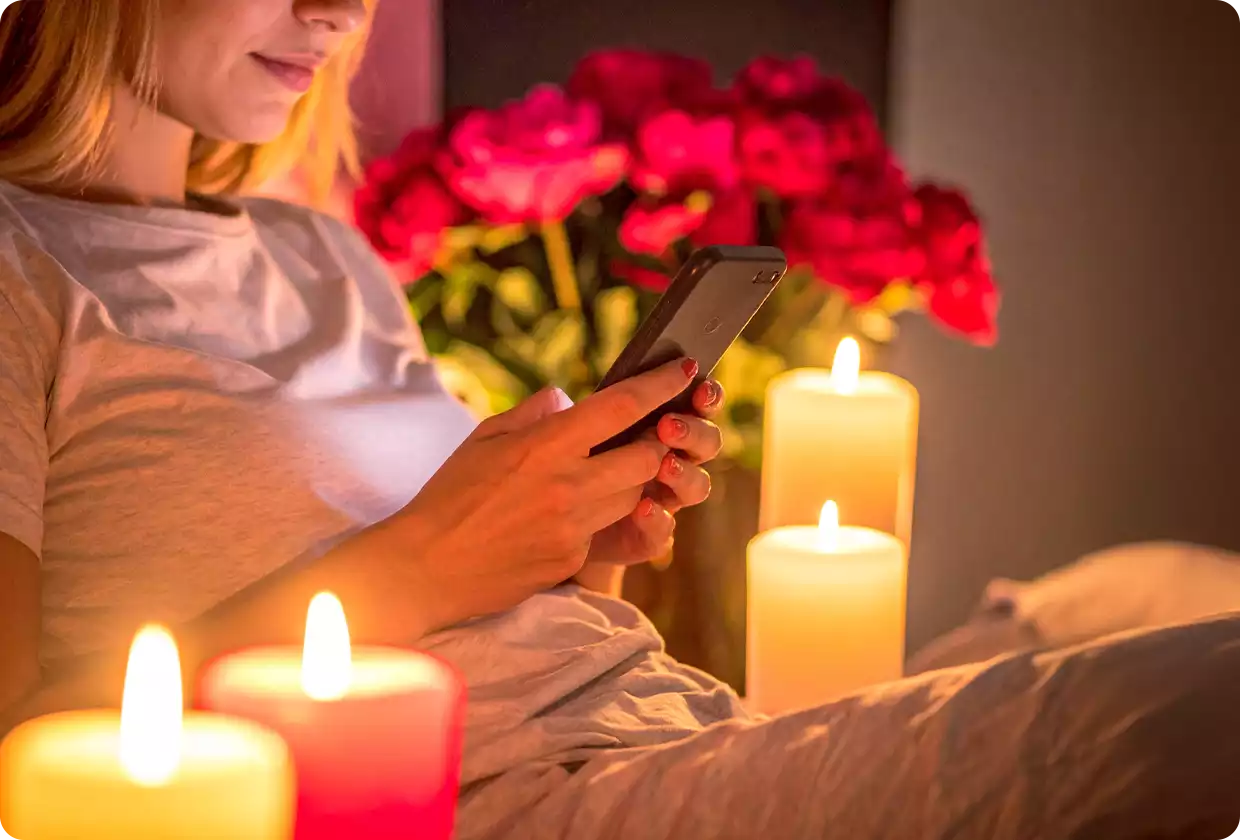 Auf Sofa sitzende Frau mit Handy in der Hand, umgeben von Kerzen und einem Rosenstrauß im Hintergrund