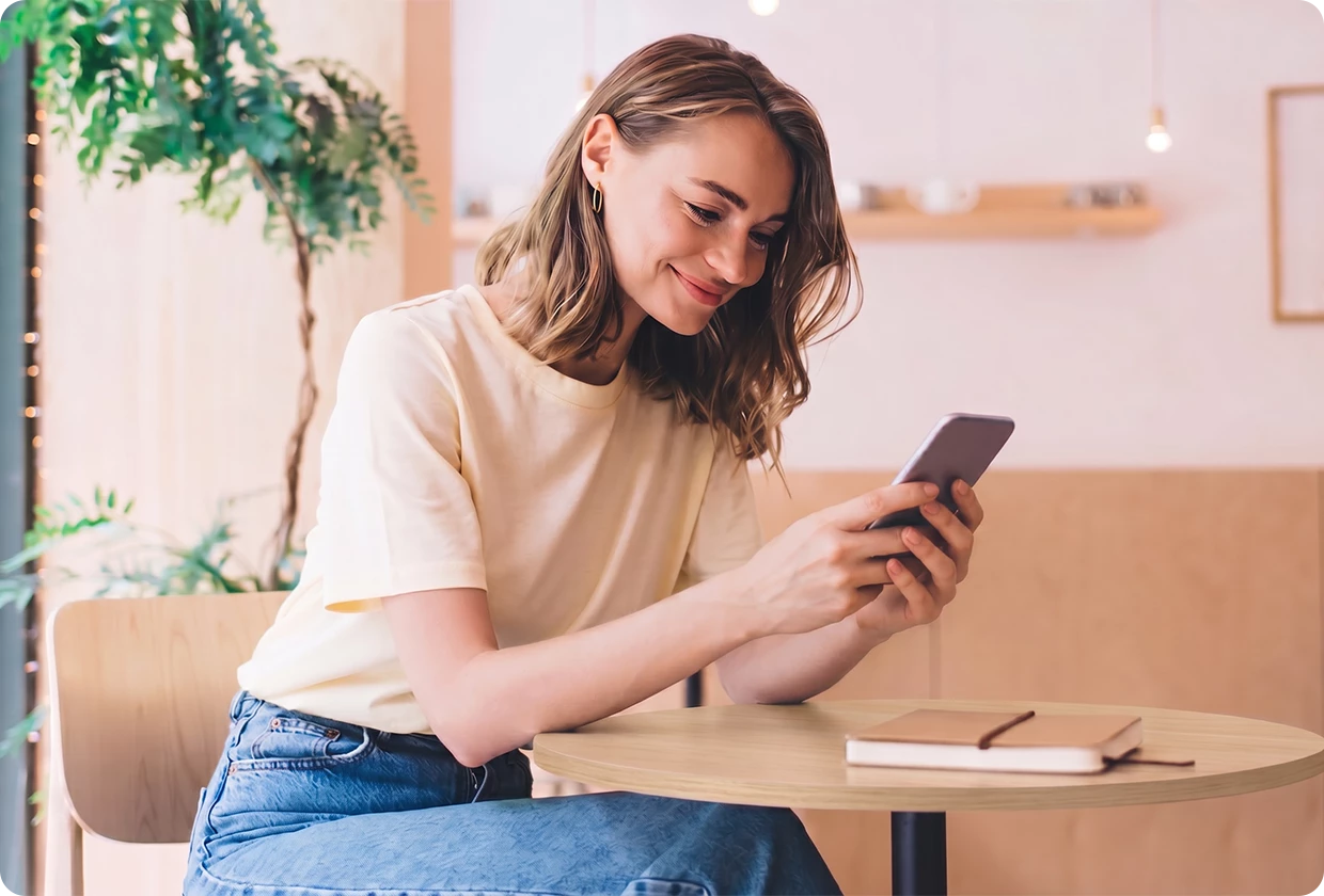 Junge Frau, die an einem Tisch sitzt und entspannt lächelnd auf ihr Handy schaut