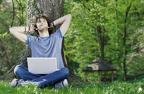Junger Mann mit Drei-Tage-Bart sitzt auf einer grünen Wiese mit einem Laptop auf dem Schoß und arbeitet