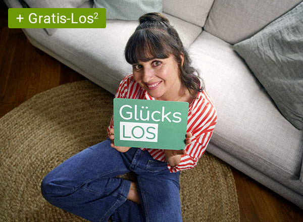 Frau mit rot-weiß gestreiftem Hemd hält rotes Smartphone vor sich, sitzt an Holztisch vor Bücherregal und Fenster mit Blick ins Grüne.