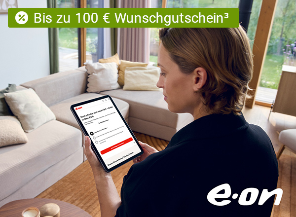 Person mit braunen Haaren hält Tablet mit E.ON-Webseite, sitzt vor hellgrauem Sofa mit Kissen in hellem Wohnzimmer.
