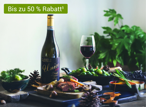 Flasche Rotwein mit goldener Kapsel und schwarzem Etikett 'Unico', daneben Weinglas mit Rotwein, grüne Trauben und Tannenzapfen auf Tisch.