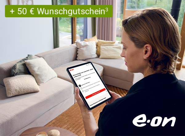 Person mit braunen Haaren hält Tablet mit E.ON-Webseite, sitzt vor hellgrauem Sofa mit Kissen in hellem Wohnzimmer.