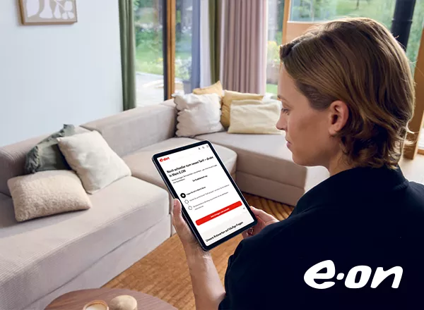 Person mit braunen Haaren hält Tablet mit E.ON-Webseite, sitzt vor hellgrauem Sofa mit Kissen in hellem Wohnzimmer.
