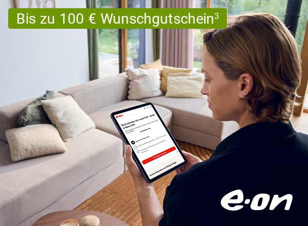 Person mit braunen Haaren hält Tablet mit E.ON-Webseite, sitzt vor hellgrauem Sofa mit Kissen in hellem Wohnzimmer.