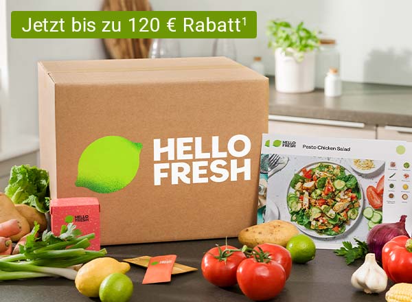 Eine HelloFresh Essensbox steht auf einem Tisch. Um die Box herum sind Kartoffeln, Paprika, Lauch, Mais, und weiteres Gemüse drapiert. An der Essensbox lehnt eine Rezeptkarte mit einem Kochrezept. Im Hintergrund sind die Umrisse einer Küchenzeile erkennbar.