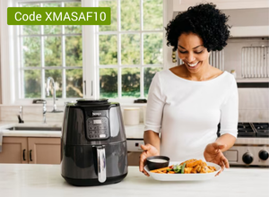 Frau in weißem Oberteil steht in moderner Küche vor schwarzer Heißluftfritteuse und hält Teller mit frittiertem Essen und Dip-Schale.