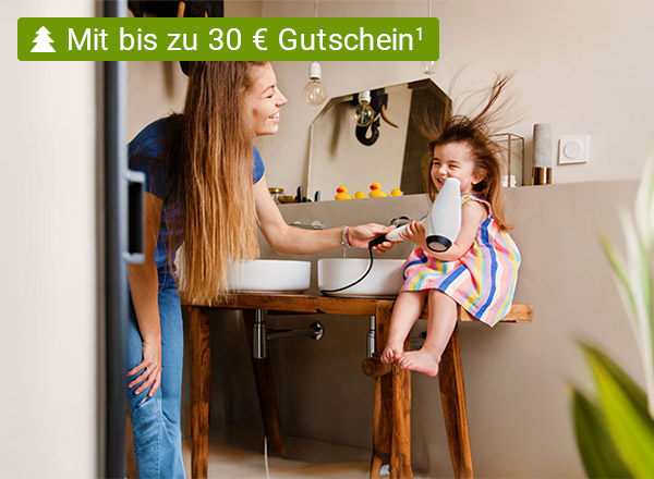 Eine Frau föhnt einem kleinen Mädchen in einem Badezimmer die Haare.  Die Haare des Mädchens sind durch den Fön zerzaust. Beide lächeln fröhlich.