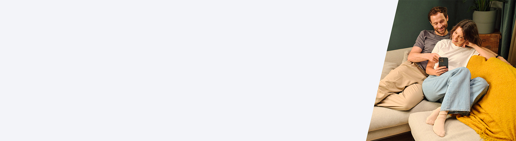 Zwei Personen sitzen entspannt auf einem Sofa, eine hält ein Smartphone, im Hintergrund steht ein Blumentopf auf einem Holzmöbel.