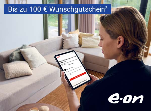 Person mit braunen Haaren hält Tablet mit E.ON-Webseite, sitzt vor hellgrauem Sofa mit Kissen in hellem Wohnzimmer.