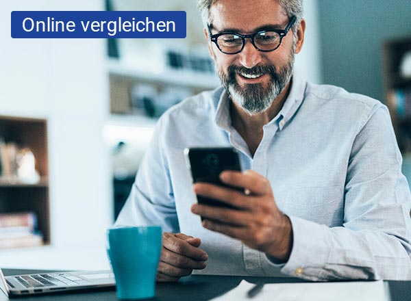 Ein Mann sitzt am Schreibtisch und schaut auf das Smartphone, welches sich in seiner Hand befindet, Auf dem Schreibtisch befindet sich ein Papierstapel, ein Kugelschreiber und eine Tasse.