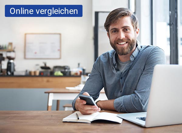 Ein junger Mann sitzt im Büro an seinem Schreibtisch. Vor ihm steht ein silbernen Laptop an dem er arbeitet. Auf dem Laptop ist ein WLAN-Zeichen in der Farbe blau und rot zu sehen.
