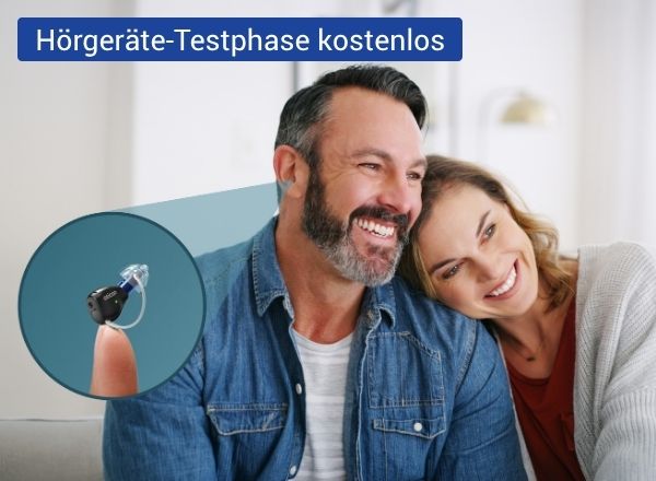 Ein lächelndes Paar sitzt eng nebeneinander in einem hellen Raum. Im Vordergrund ist zusätzlich ein kleines Hörgerät zu sehen, das auf einem Finger präsentiert wird.