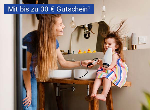 Eine Frau föhnt einem kleinen Mädchen in einem Badezimmer die Haare. Die Haare des Mädchens sind durch den Fön zerzaust. Beide lächeln fröhlich. Das Bild enthält eine Abbildung eines BestChoice-Gutscheins mit 20 Euro Gutscheinwert.