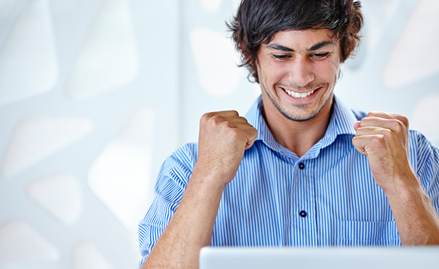 An ecstatic young businessman wearing a blue shirt sitting in front of his laptop