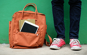 A backpack containing a tablet computer on the ground next to a kid’s feet wearing sneakers