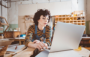 Woman wearing overalls in a carpentry workshop looks at a laptop with a serious expression on her face