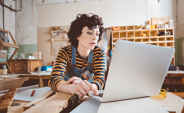 Woman wearing overalls in a carpentry workshop looks at a laptop with a serious expression on her face. 