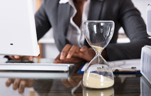 Hourglass on a desk with sand falling as a person works at a computer in the background.