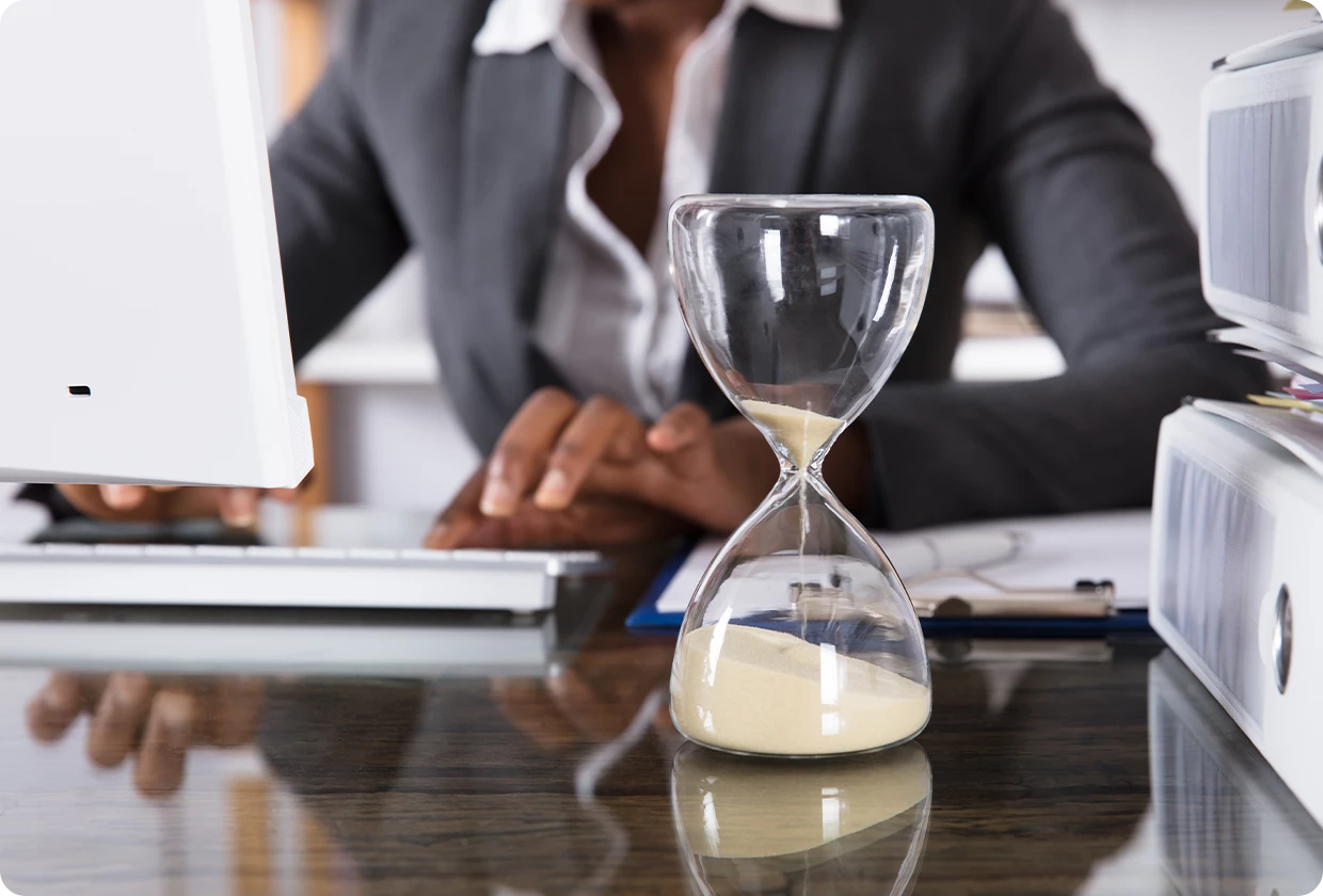 Hourglass on a desk with sand falling as a person works at a computer in the background.