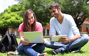 Two students sitting on the grass outdoors, smiling while using a laptop and a tablet together.