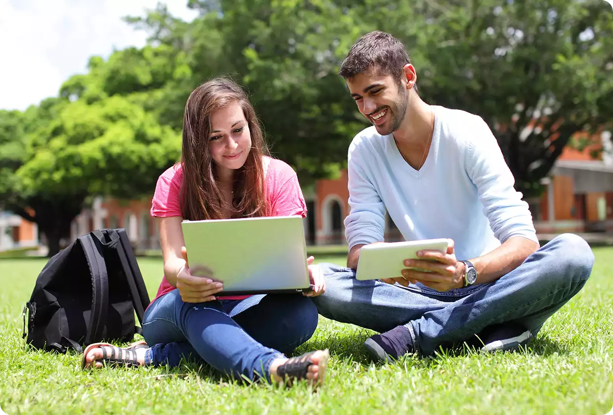 Two students sitting on the grass outdoors, smiling while using a laptop and a tablet together.