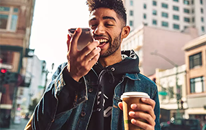 A young man records a voice message on his smartphone while holding a coffee.