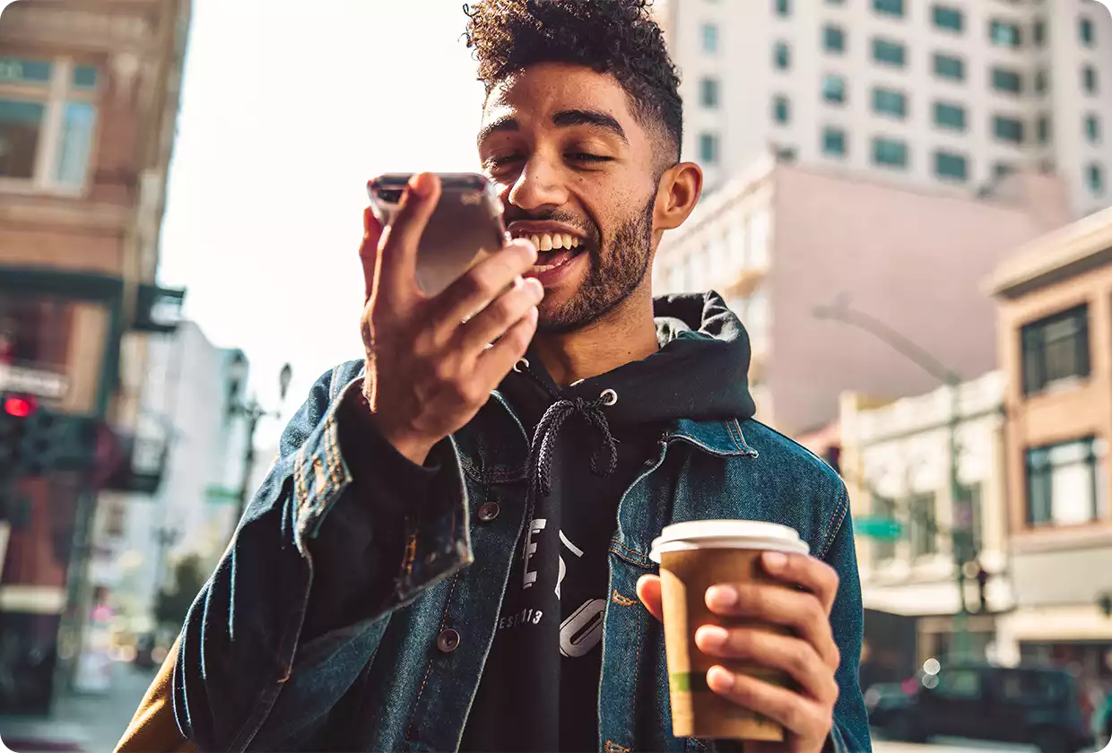 A young man records a voice message on his smartphone while holding a coffee.