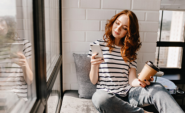 Young woman wearing striped shirt holds smartphone in one hand and coffee cup in other