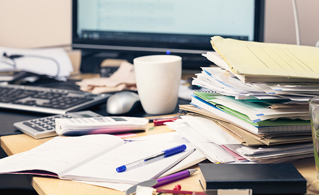 Stack of files and documents on messy desktop, PC monitor in background