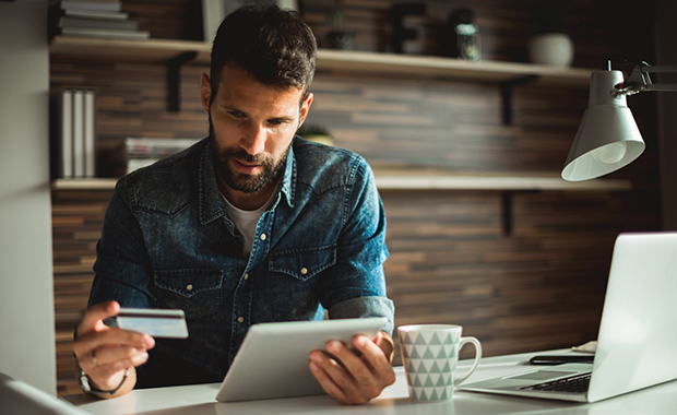 Bearded man at desk holding credit card and looking at tablet computer