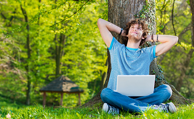 Smiling young man with laptop sits on grass leaning against tree trunk