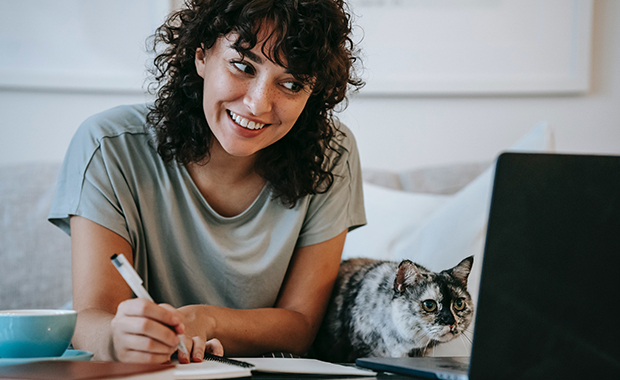 Smiling woman and cat sit on couch looking at laptop