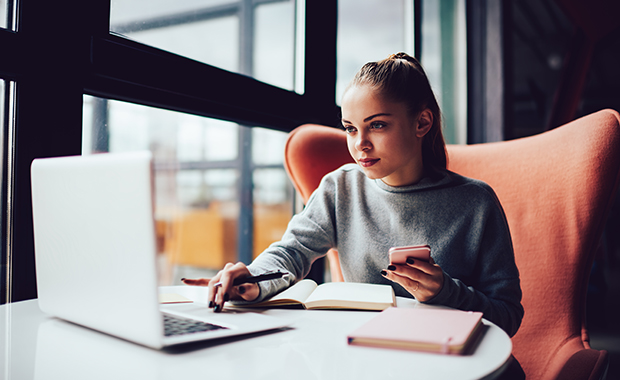 Woman sitting in café looking at laptop while holding smartphone