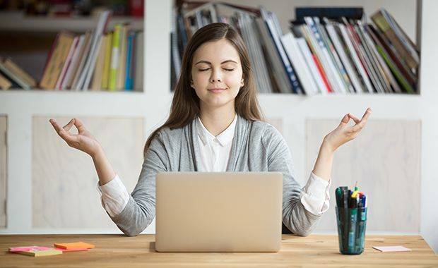 Smiling women sits in front of laptop with eyes closed making the Gyan mudra meditation gesture with each hand