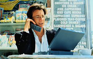 Man sits in café working on laptop and speaking on cell phone. The devices date the scene to the late 1990s