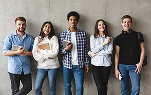 Group of students leaning on a gray wall and smiling at the camera.