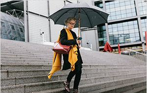 An older woman walks through the rain holding an open umbrella while looking at her smartphone.