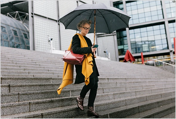 An older woman walks through the rain holding an open umbrella while looking at her smartphone.