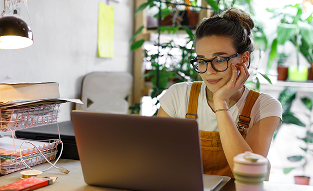 Young woman sits at desk in home office and looks at laptop