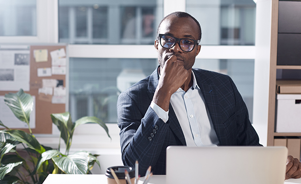 Businessman wearing suit and glasses sits thinking at desk with laptop 