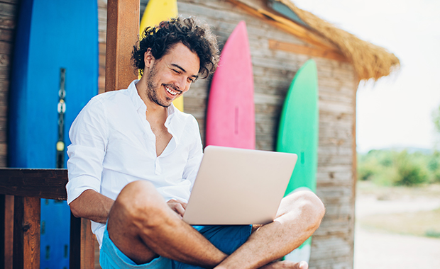 Smiling man sits cross-legged with laptop leaning against colorful surf boards