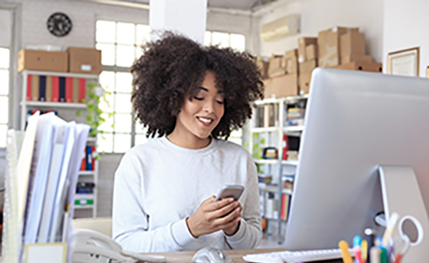 Woman sits in front of computer screen in home office looking at phone