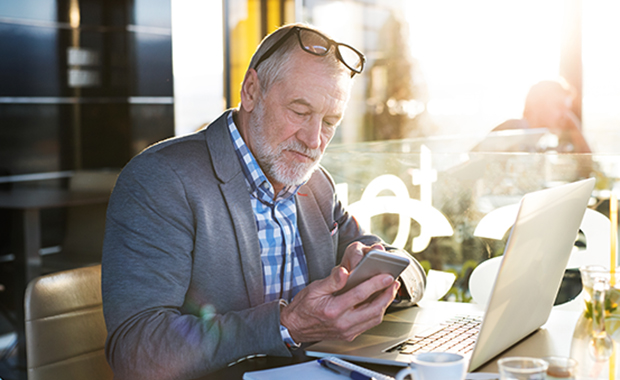 Senior businessman sits at cafe table working on laptop and looking at phone