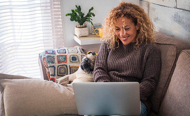 Woman and pug dog sit on couch looking at laptop