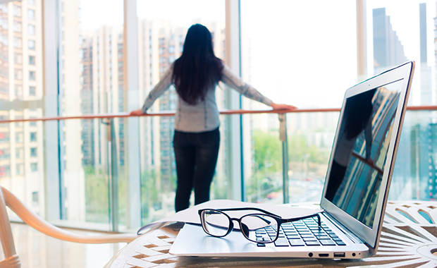 Open laptop and glasses on table with woman looking out window in background