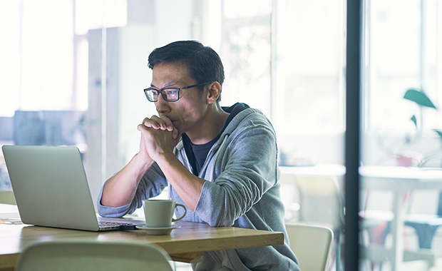 Man in hoodie sits in office looking at laptop with serious expression on face